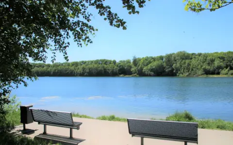 Zwei Bänke laden zum Verweilen am Ufer des halener Badesees ein. Bäume säumen das klare Wasser unter blauem Himmel.