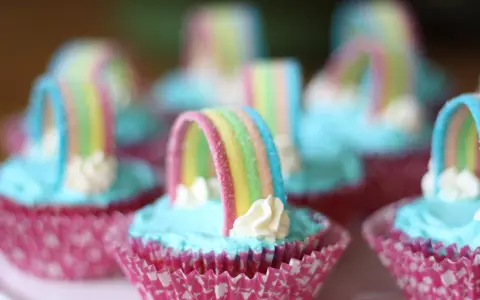 Sju cupcakes med ljusblå regnbågs frosting och godisregnbågar. Rosa muffinsformar med mönster.