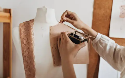 Mani che lavorano su un manichino, creando un modello di cartone. Un corso di modellistica in pieno svolgimento, con pizzi e accessori da cucito a portata di mano.