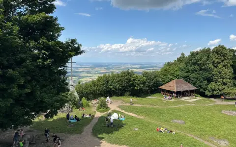 Ludzie odpoczywają na trawie i spacerują po szlakach na górze Ślęża. W oddali widać rozległą panoramę.