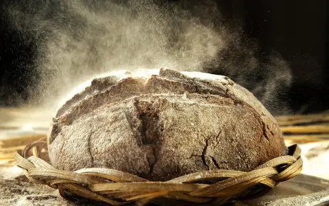 Un pain au seigle rustique, saupoudré de farine, repose dans un panier en osier. La poussière de farine danse dans la lumière, créant une atmosphère magique.