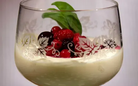 En elegant glas med vit chokladmousse, toppad med färska bär och ett grönt blad. Glaset har en vacker dekor av löv.