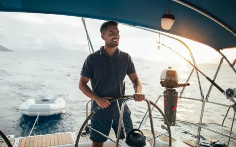 Un homme souriant est au gouvernail, prêt à barrer un bateau. Le soleil brille sur l'eau calme.
