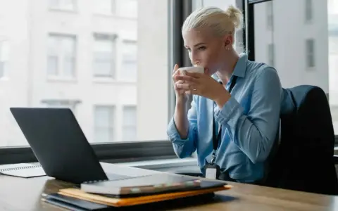 Donna bionda sorseggia caffè mentre lavora al laptop, cercando concorsi psicologi non scaduti.