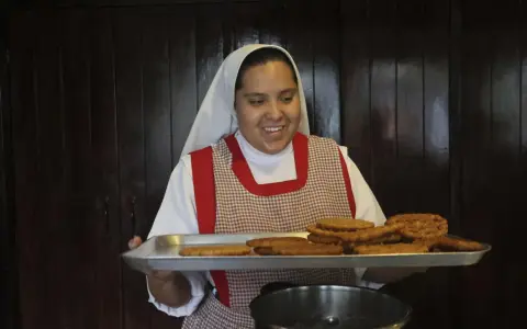 A smiling nun in a white habit and checkered apron holds a tray of fried pastries, perhaps answering the question: what do nuns eat for breakfast?