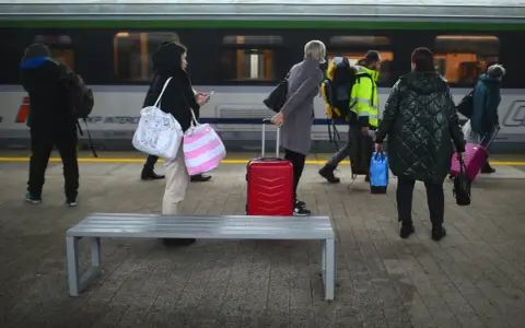 Ludzie z bagażami czekają na peronie PKP Intercity. Czerwona walizka stoi obok ławki.