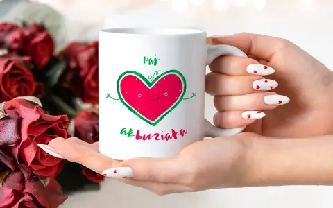 Biały kubek z zabawnym sercem-arbuzem i napisem "Daj buziaka", trzymany w dłoniach z paznokciami w serduszka, na tle czerwonych róż.