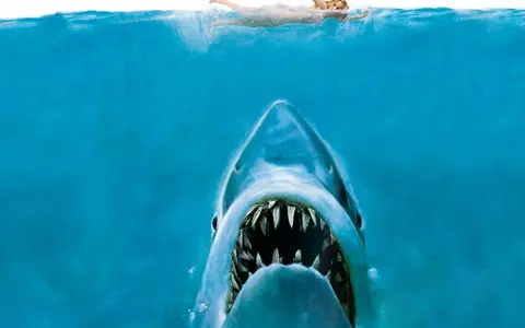 Un tiburón emerge de las profundidades, con la boca abierta, mientras una nadadora se acerca a la superficie. La icónica banda sonora de tiburón resuena.