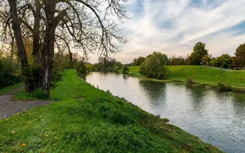 Spokojna rzeka na u, otoczona zielonymi brzegami i drzewami. W oddali widać ścieżkę i niebo z chmurami.