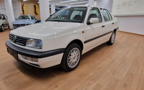 Biały Volkswagen Vento z lat 90. stoi na drewnianej podłodze w salonie samochodowym.