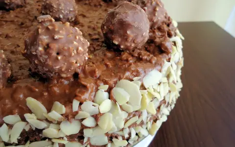 Pyszny tort Ferrero Rocher, oblany czekoladą i obsypany płatkami migdałów, z całymi czekoladkami na wierzchu.