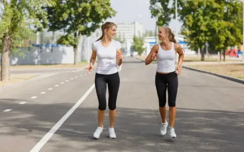 Due donne sorridono mentre praticano diversi tipi di camminata sportiva su una strada asfaltata, con alberi e edifici sullo sfondo.
