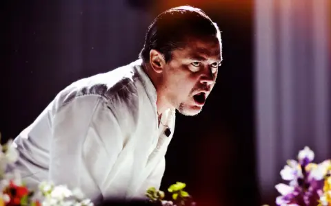 Mike Patton heute, mit weit aufgerissenem Mund und intensivem Blick, trägt ein weißes Hemd. Blumen im Vordergrund.