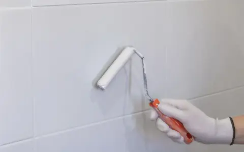 Mano enguantada pintando con rodillo blanco sobre azulejos grises. Renovando el aspecto de los tubos calefacción.