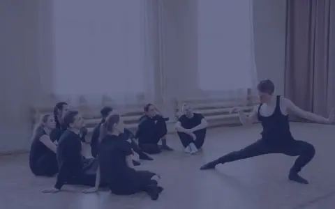 Choreograf demonstruje ruchy grupie tancerzy siedzących na podłodze.