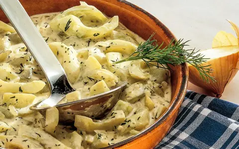 Cremiges Schmorgurken Rezept mit Dill in einer Tonschale, daneben eine Zwiebel und ein Löffel.