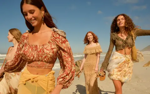 Quattro donne con abiti in stile boho passeggiano su una spiaggia sabbiosa con il mare sullo sfondo.