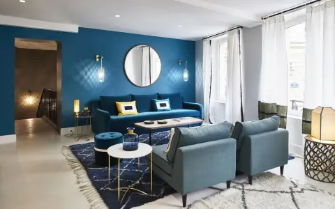 Salon moderne avec mur bleu canard, canapé velours, fauteuils et tapis assortis. Le jaune vif des coussins dynamise l'ensemble.
