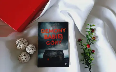 Książka "Demony Babiej Góry" Ireny Małysy, leżąca na białym jedwabiu, obok czerwonego pudełka i ozdobnych kulek. Historia, która zabiera daleko od Babiej Góry.