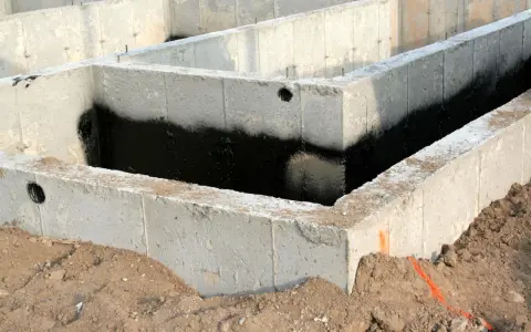 Surowe ściany fundamentowe z betonu, częściowo pokryte czarną izolacją przeciwwilgociową, otoczone ziemią.