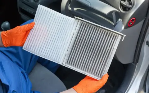 Ręce w pomarańczowych rękawiczkach pokazują brudny filtr kabinowy Toyoty Yaris, gotowy do wymiany.