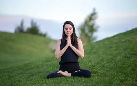 Kvinna i meditationsposition på en grön kulle, med slutna ögon och händerna i bön.