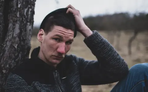 Quebonafide w czapce i swetrze, oparty o drzewo. W tle jesienny krajobraz. Tekst "żadnych zmartwień" pasuje do nastroju.