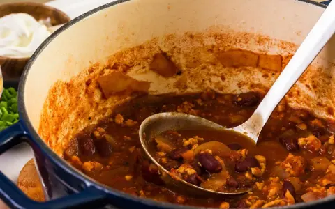 Gęste, aromatyczne chili con carne w garnku, z widoczną łyżką nabierającą porcję. Obok miseczka ze śmietaną.