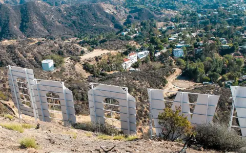 Widok zza gigantycznych liter znaku Hollywood. W oddali widać wzgórza z domami i drogami.