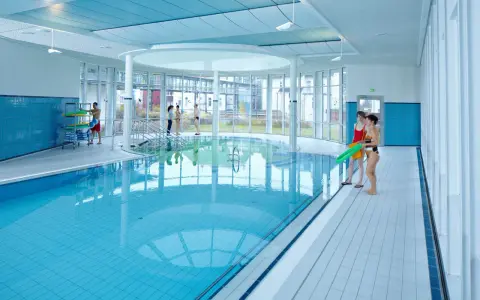 Menschen in Badekleidung am Beckenrand eines Hallenbades mit türkisfarbenem Wasser.