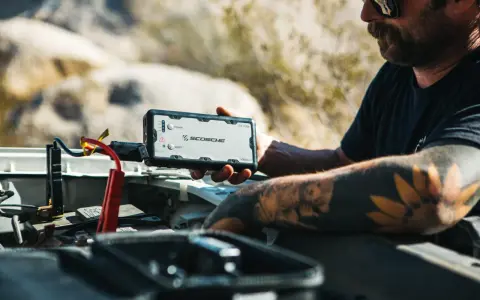 Man with tattoos demonstrates how to charge a jump starter by connecting it to a car battery in a desert landscape.