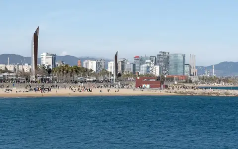 Ludzie odpoczywają na plaży, a w tle widać nowoczesne budynki Barcelony. To miasto leży nad Morzem Śródziemnym.