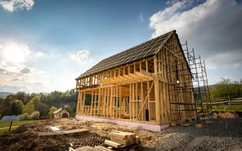 Widok na budowę domu szkieletowego. Drewniana konstrukcja z widocznymi belkami i stropami, otoczona rusztowaniami. W tle zielone wzgórza i błękitne niebo.
