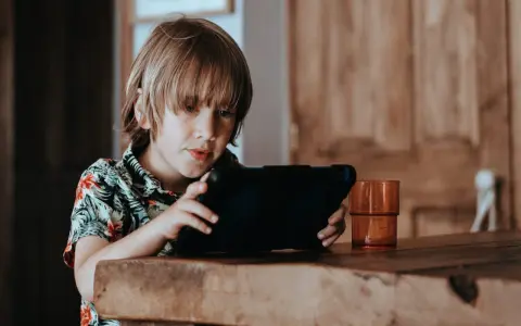 Chłopiec w hawajskiej koszuli ogląda krótkie filmy na tablecie, siedząc przy drewnianym stole.