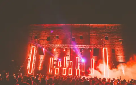 Folla in delirio al **mood festival**, illuminata da luci al neon rosse e fumo denso. La scena è un'esplosione di energia.