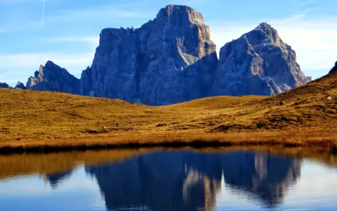 Riflessi maestosi delle Dolomiti nel lago delle Baste, con un prato dorato che separa le acque dalle vette imponenti.