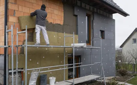 Pracownik na rusztowaniu montuje wełnę mineralną na ścianie domu. Brak cokołu na elewacji, trwa ocieplanie.