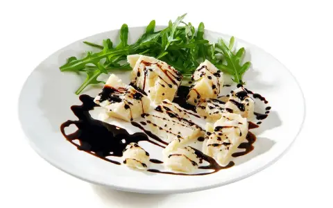 Kawałki parmezanu polane gęstą glazurą balsamiczną, podane z rukolą na białym talerzu.