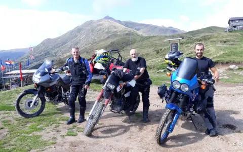 Tre motociclisti con moto da enduro posano in cima al passo San Lucio, con montagne sullo sfondo.