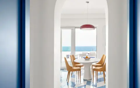 Interno di una casa stile mare con pavimento a scacchi blu e bianchi, tavolo rotondo con sedie arancioni e vista sul mare.
