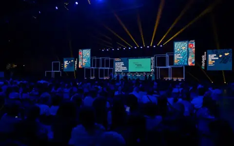 Un grande pubblico partecipa a un meeting per l'amicizia fra i popoli, illuminato da fasci di luce gialla su un palco con schermi.