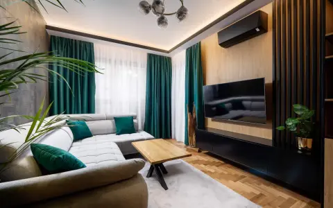 Nowoczesny salon z szarą kanapą i zielonymi poduszkami. Po prawej stronie ściana ozdobiona pionowymi lamelami w narożniku, na niej telewizor.