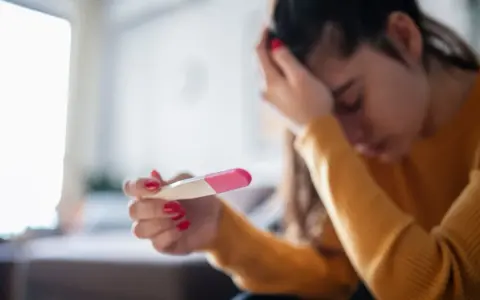 Kobieta z opuszczoną głową trzyma test ciążowy. Jej dłoń z czerwonym lakierem do paznokci zaciska się na teście. Tokofobia może wywoływać takie emocje.