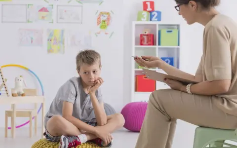 Chłopiec siedzi na poduszce, zastanawiając się, jakie pytania zadaje psycholog dziecku. W tle rysunki dzieci i kolorowe zabawki.