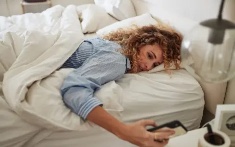 Kobieta z kręconymi włosami leży w łóżku, sięgając po telefon. Czyżby 14:44 znaczenie miało dla jej poranka?