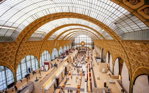 Interior del museo D'Orsay, con su impresionante techo abovedado de cristal y arcos dorados. Gente pasea entre esculturas y exposiciones.