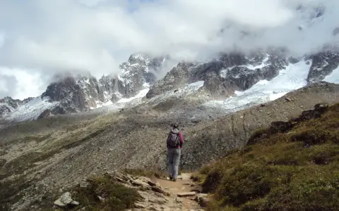 Osoba z plecakiem i kijkami idzie ścieżką w górach. W tle majestatyczne szczyty Alp, częściowo pokryte śniegiem i chmurami. Wspaniały widok na trekking w alpach.