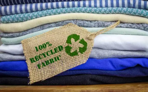 Pile di tessuti ecologici colorati, con un'etichetta che indica "100% Recycled Fabric" e il simbolo del riciclo.
