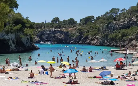En livlig strand vid Cala Pi, med folk som badar i det turkosa vattnet och solar på sanden under parasoller.