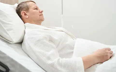 Mężczyzna w szpitalnym łóżku, w białym szlafroku, po operacji obniżenia pęcherza moczowego.